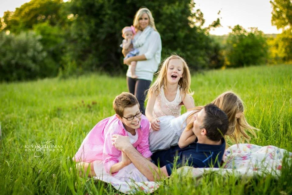 Family playing on a picnic blanket in a grassy field with trees in the background, mother holding a baby, father lying on ground with children playing around him.