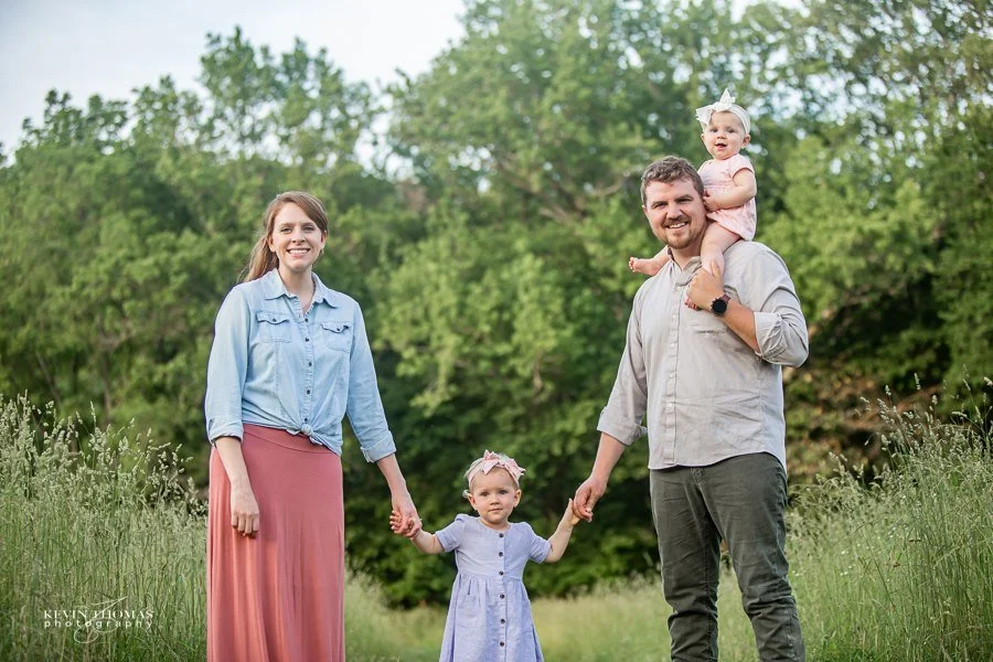A family of four holding hands in a green field with trees in the background. The mother has long brown hair and is wearing a light blue denim shirt and a long pink skirt. The father has short brown hair and a beard, wearing a light gray shirt and da