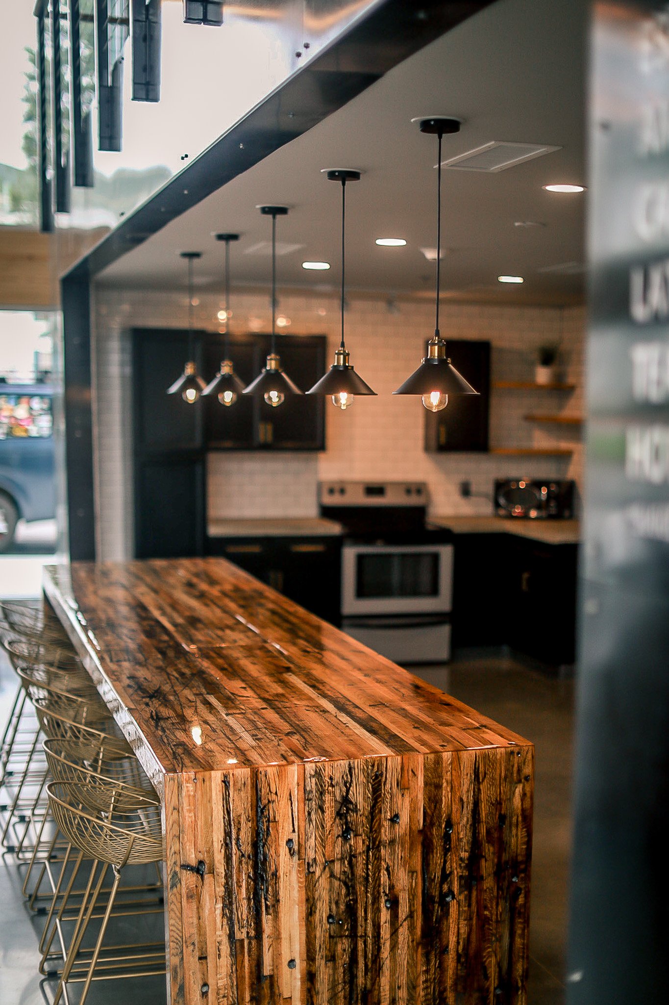 Interior of a modern kitchen with a wooden bar counter, black cabinets, stainless steel appliances, open shelves, and hanging pendant lights.