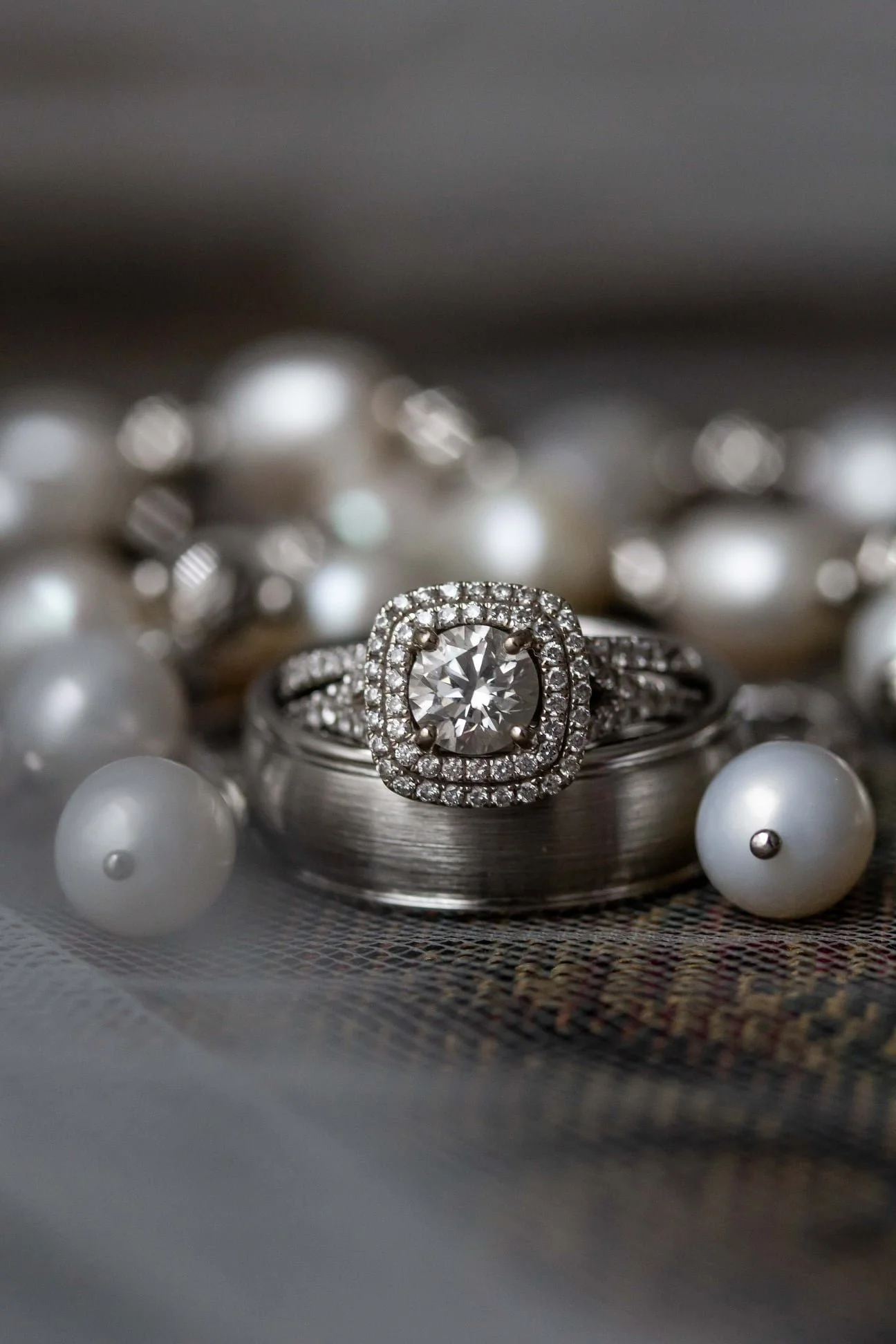 Close-up of a diamond engagement ring with a halo setting, placed on top of a wedding band, surrounded by scattered pearls.