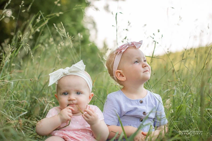 Two young children sitting in a grassy field, one holding a small stick and the other looking up at the sky, both wearing headbands and casual clothing.