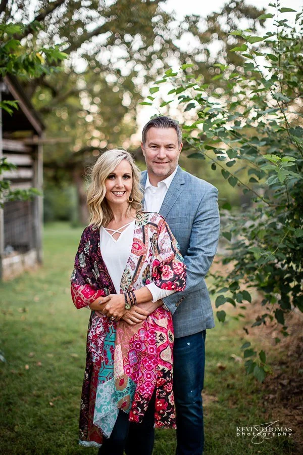 A smiling couple standing outdoors surrounded by trees, with the woman wearing a colorful printed coat and the man in a blue blazer.