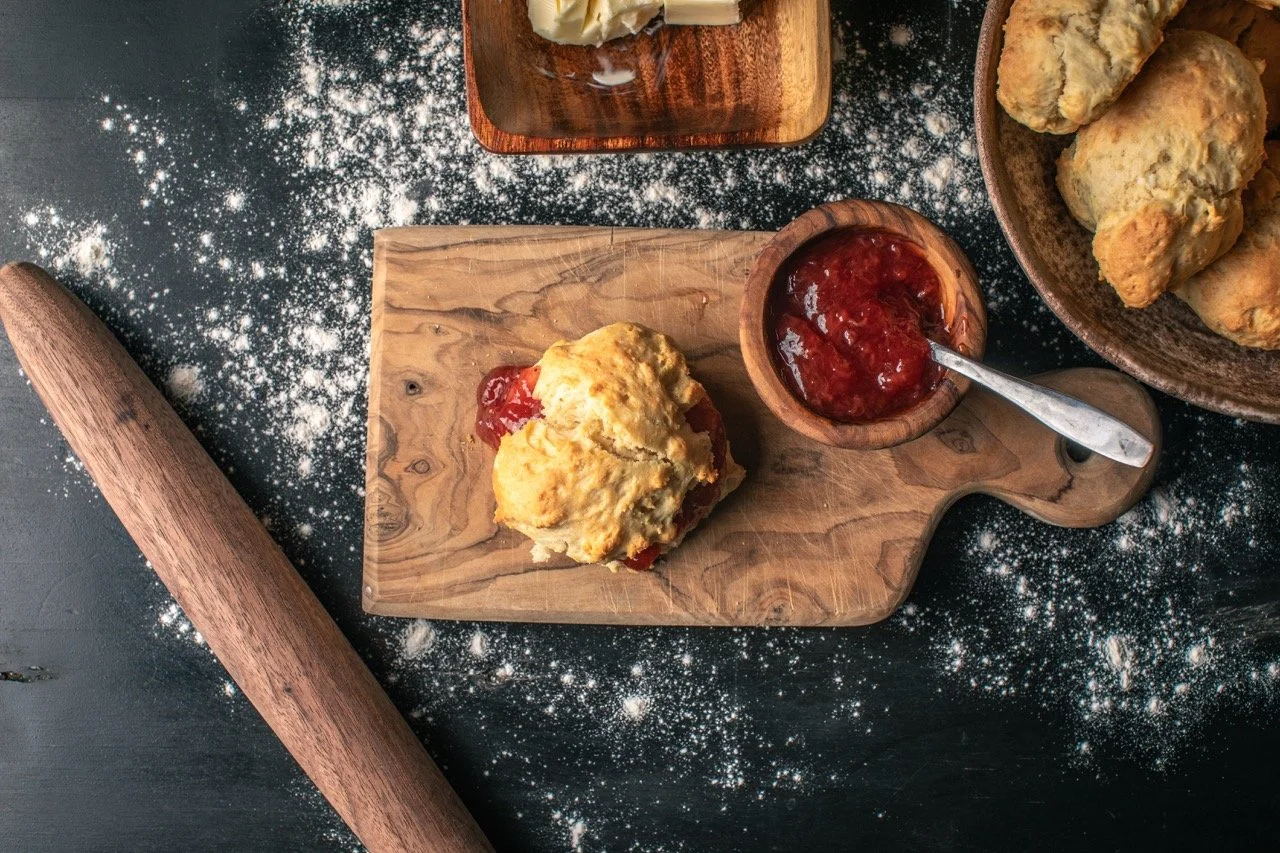 A scone with red jam on a wooden cutting board next to a small bowl of jam with a spoon, a bowl of other scones, a rolling pin, and scattered flour on a dark surface.