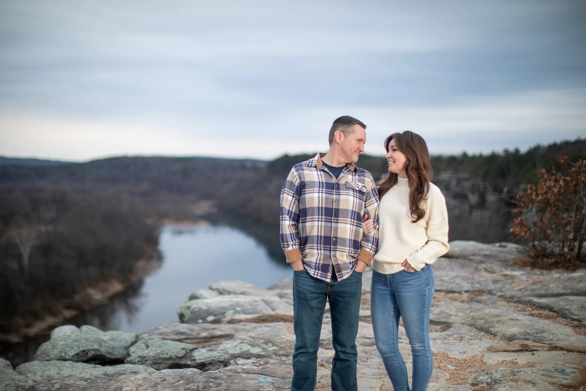 A man and woman standing close together on a rocky ledge outdoors, smiling and looking at each other with a river and wooded landscape in the background during overcast weather.