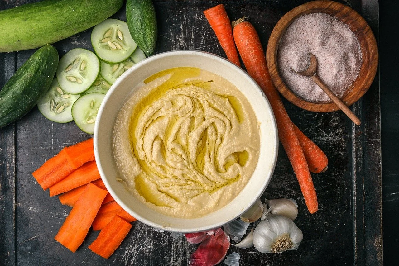A bowl of hummus with a swirl of olive oil on top, surrounded by fresh vegetables including cucumbers, carrots, garlic, and salt on a dark textured surface.