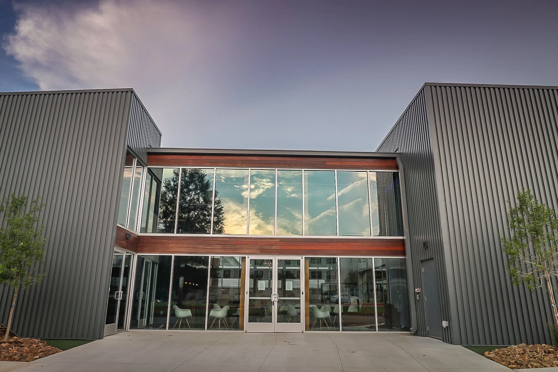 Modern building with large glass windows reflecting a sunset sky and trees, flanked by gray metal siding and surrounded by small trees and landscaping.