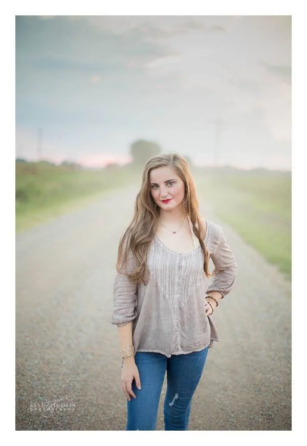 A young woman standing on a gravel road in a rural area with grassy fields and power lines in the background during daylight. She has long brown hair, is wearing a beige blouse, blue jeans, and is looking at the camera with a slight smile.