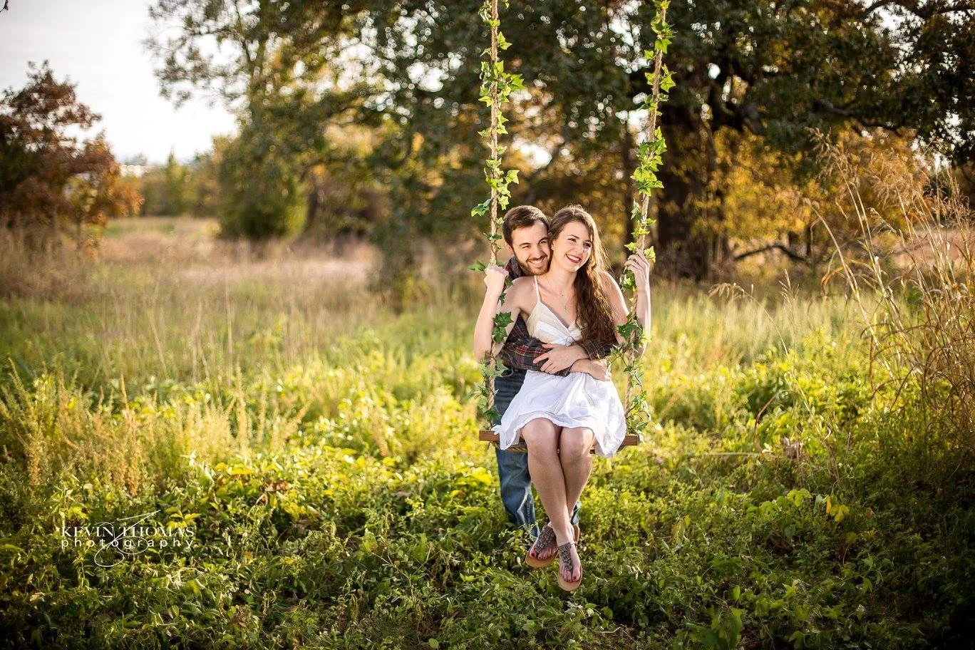 A smiling couple sitting on a swing decorated with green leaves in a field during late afternoon, surrounded by tall grass and trees with autumn foliage.