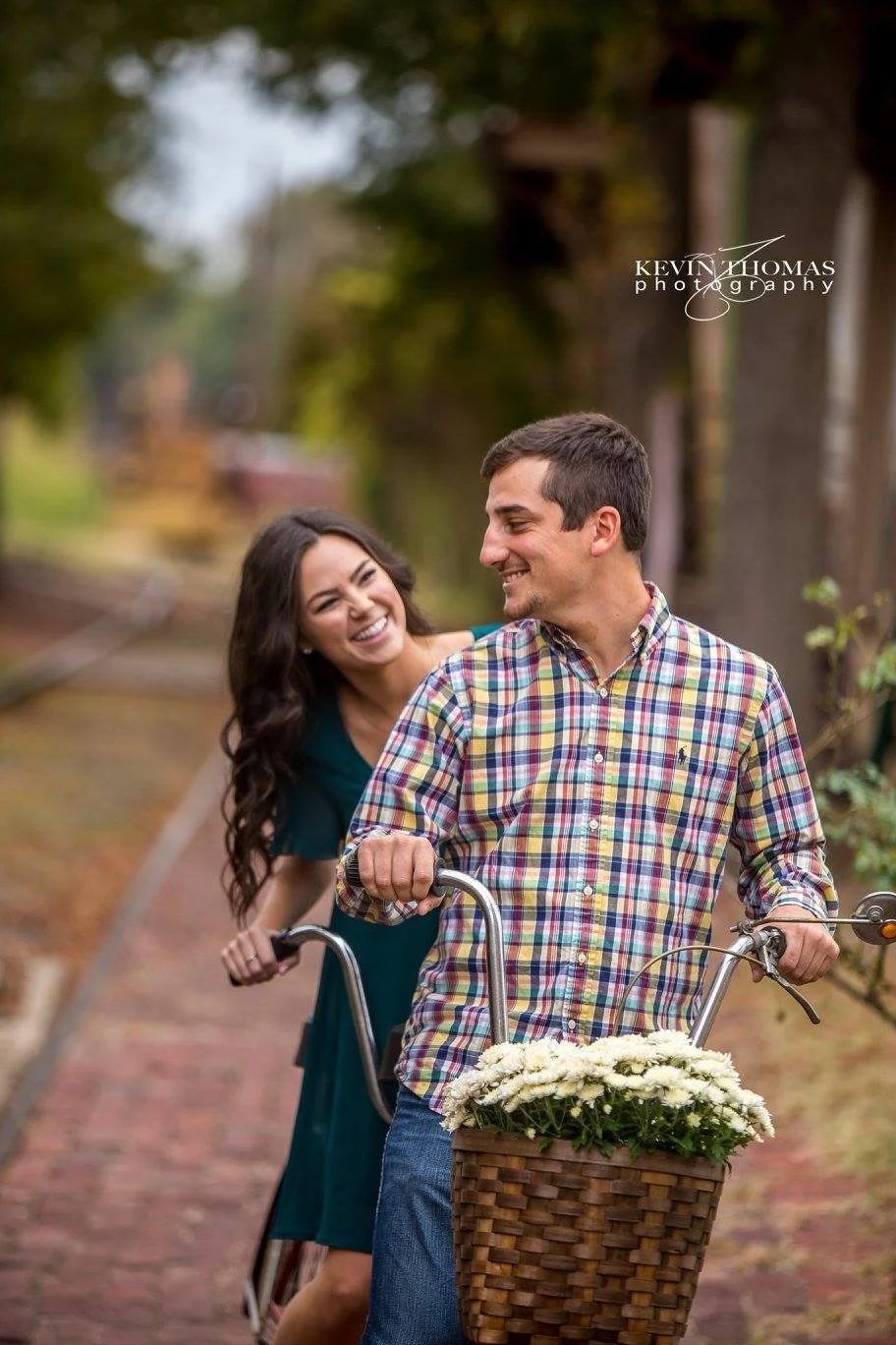 A smiling woman riding behind a man on a bicycle with a basket of white flowers, on a brick path in a park or garden.