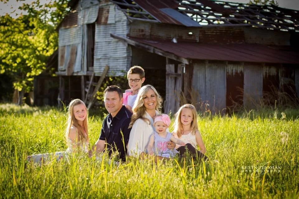 A family of six sitting and standing in a grassy field near a rustic barn, smiling at the camera on a sunny day.