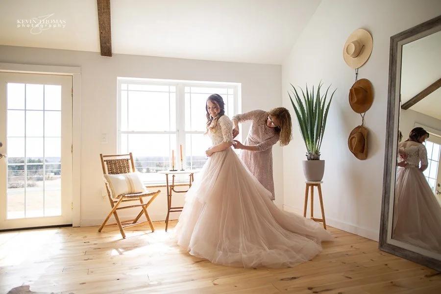 A woman in a wedding dress being helped by another woman in a light pink dress to adjust her gown inside a bright, minimalist room with large windows, a potted plant, two hats on the wall, and a mirror reflecting both women.