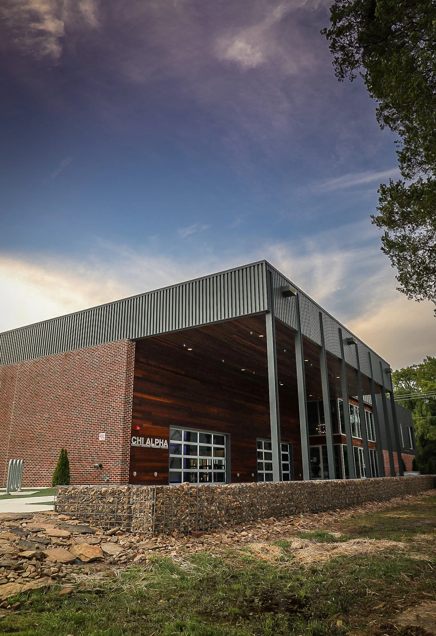 Modern industrial-style building with brick and metal exterior, large glass garage doors, and a wooden accent wall. The building has a sign that reads "Chi Alpha" outside. The sky is partly cloudy and there are trees nearby.