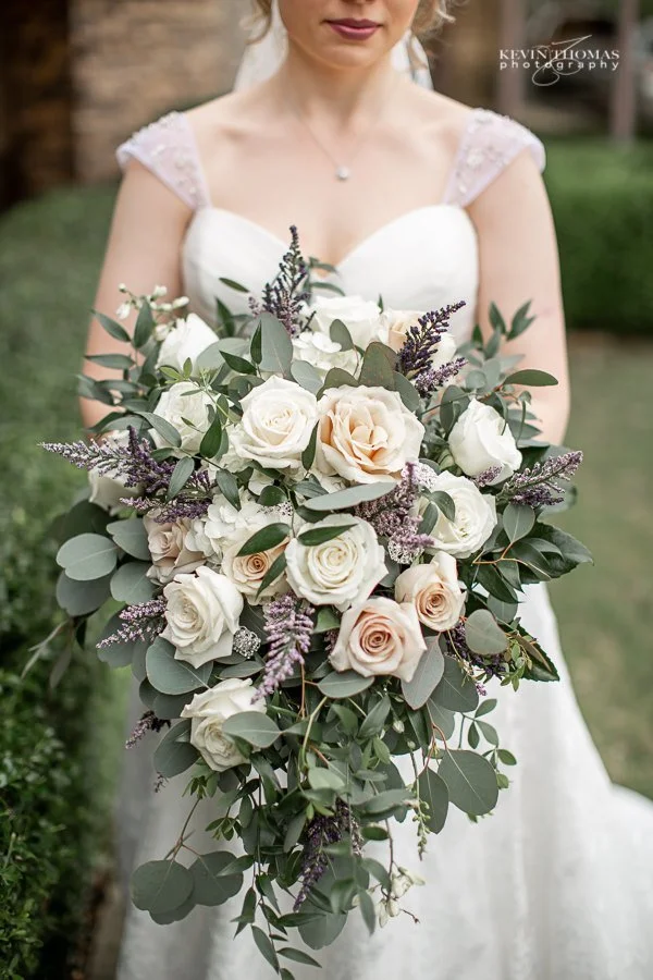 A bride in a white wedding dress holding a large bouquet of white and blush roses, surrounded by greenery and small purple flowers.