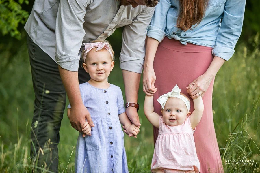 A family of four holding hands outdoors in a grassy area, with a man, woman, and two young girls in pastel dresses and headbands.
