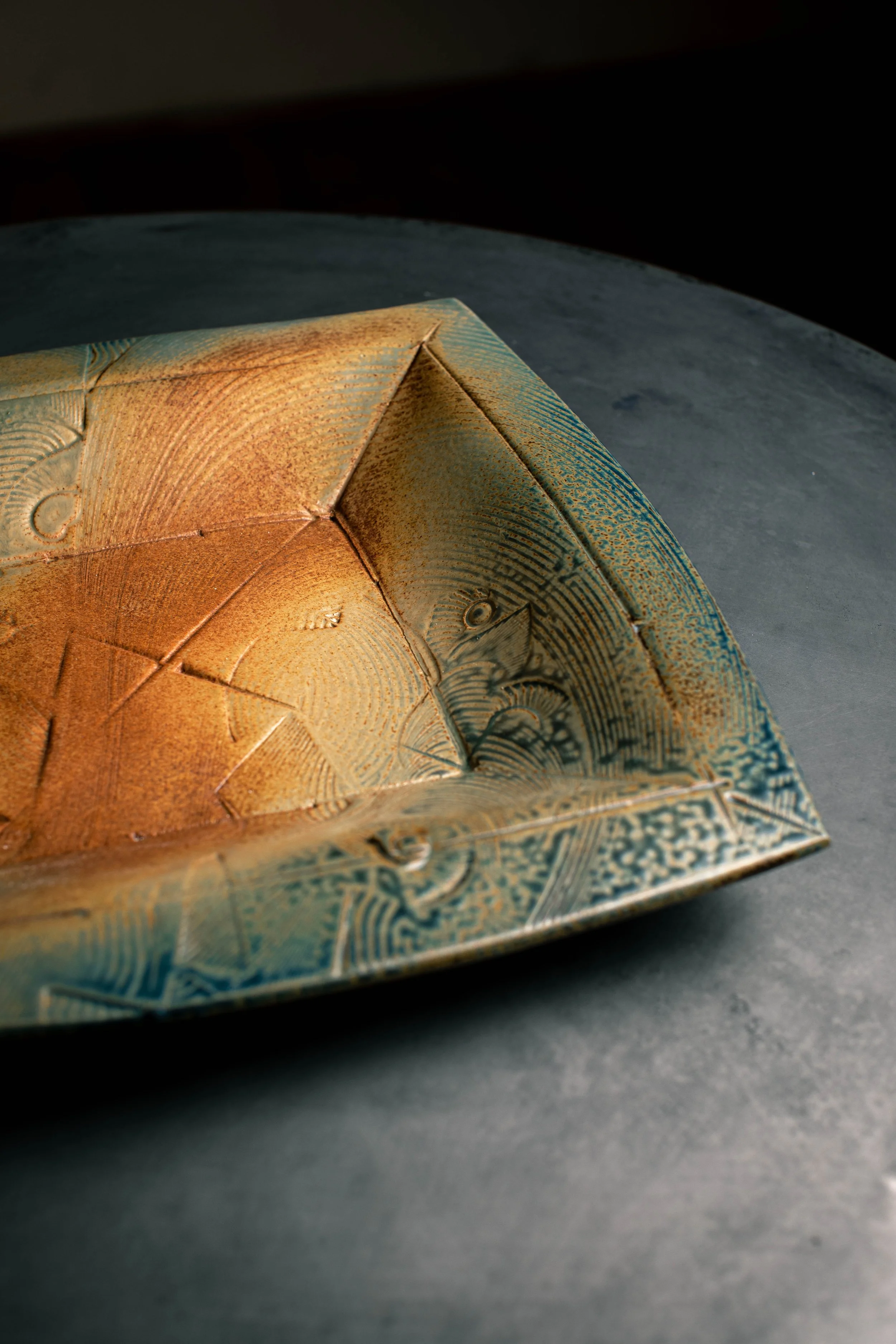 Close-up of an artistic, textured square bowl with intricate geometric patterns in shades of gold, green, and blue on a dark surface.