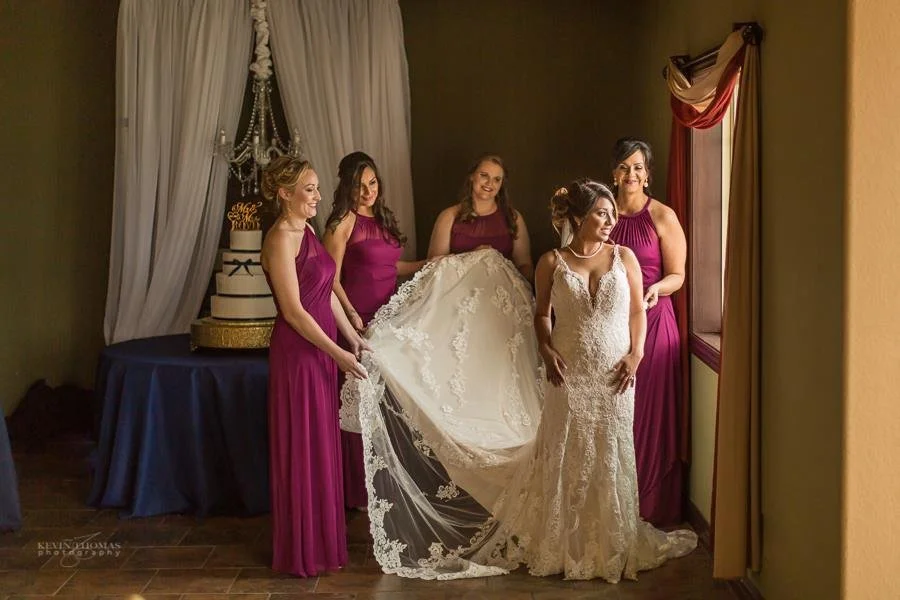 A bride in a white lace wedding gown standing with five women in magenta bridesmaid dresses, holding her train, in a room with a wedding cake on a table and decorated curtains.