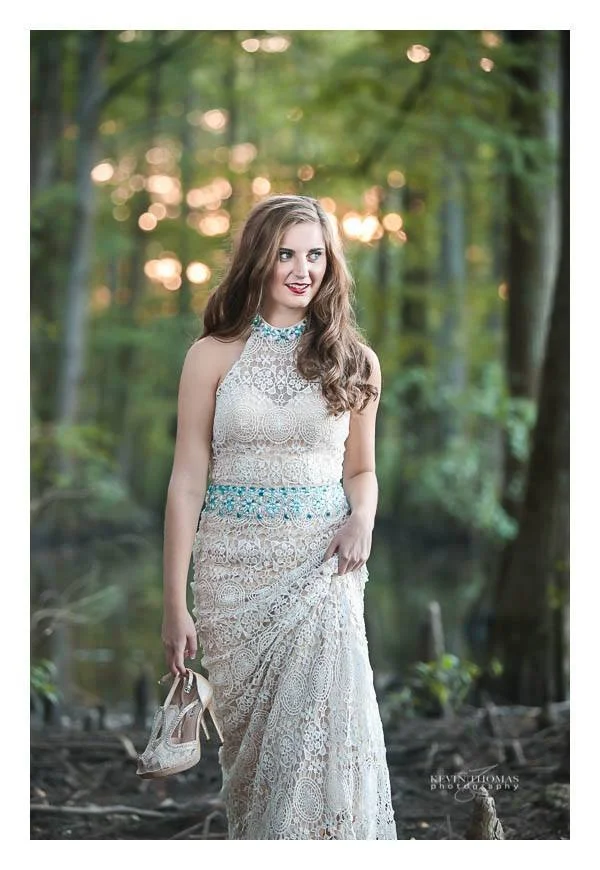 A woman wearing a lace dress holding high heels in a forest with sunlight filtering through trees.