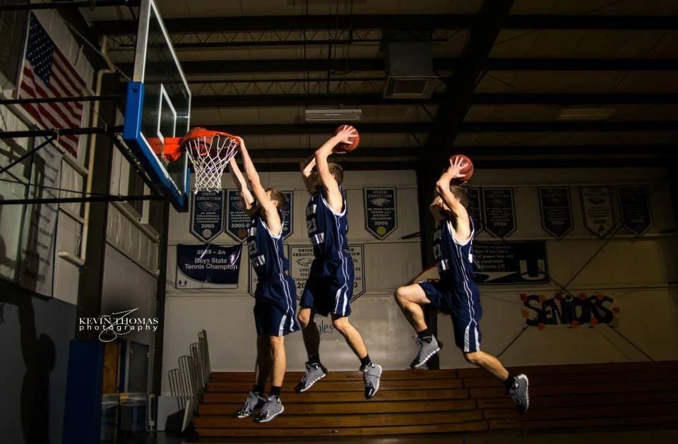 Three young male basketball players in blue uniforms jumping simultaneously to dunk or block a basket during a game or practice in a gymnasium.