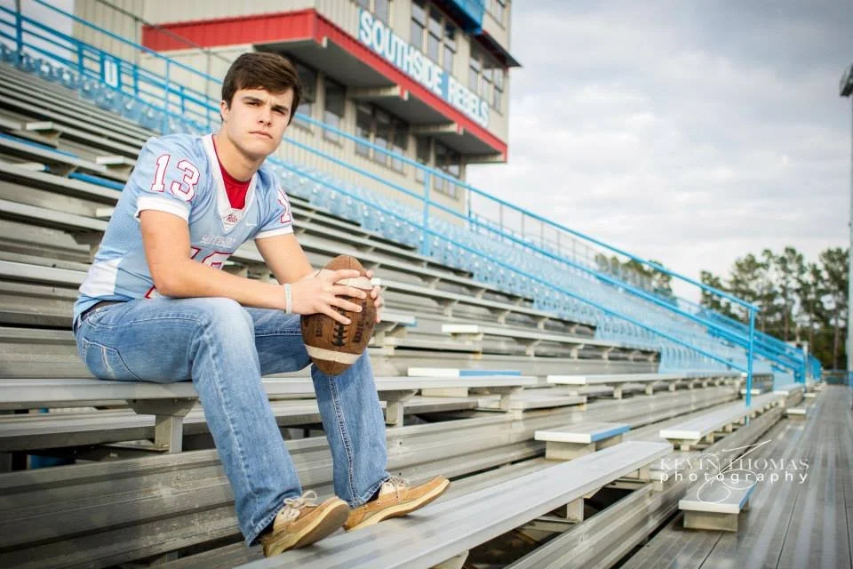 Young man sitting on bleachers at a sports stadium, holding a football, wearing a sports jersey, jeans, and sneakers, with a serious expression.