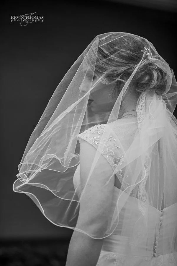 Black and white photo of a bride with her head bowed, wearing a wedding dress with pearl embellishments and a veil covering her face and shoulders.