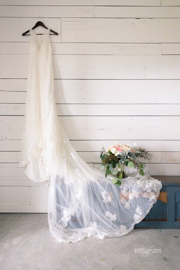 A white wedding dress hanging on a black hanger displayed against a white wooden wall, with a bouquet of flowers and lace fabric draped over a small blue and brown wooden bench nearby.