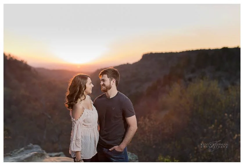 A young couple standing on a rocky ledge during sunset with a canyon in the background, smiling at each other.