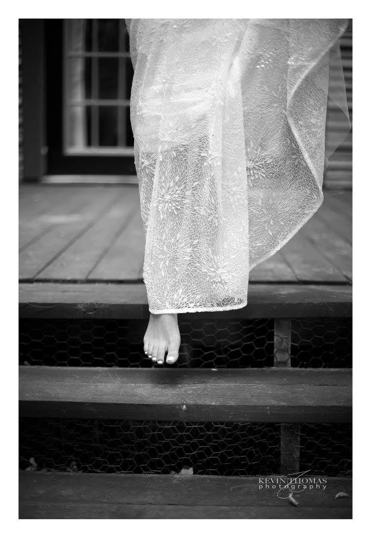 Close-up of a single foot hanging off the edge of a wooden porch, with sheer fabric draped over the leg, with a window and siding in the background.