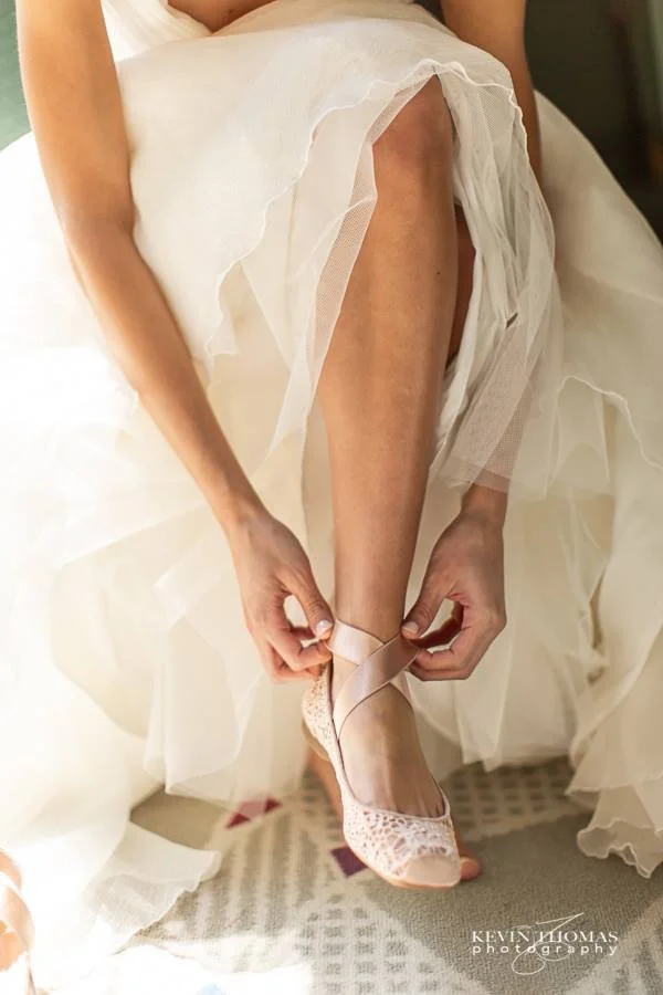 A woman in a wedding dress is putting on a pink lace high-heeled shoe on her foot.