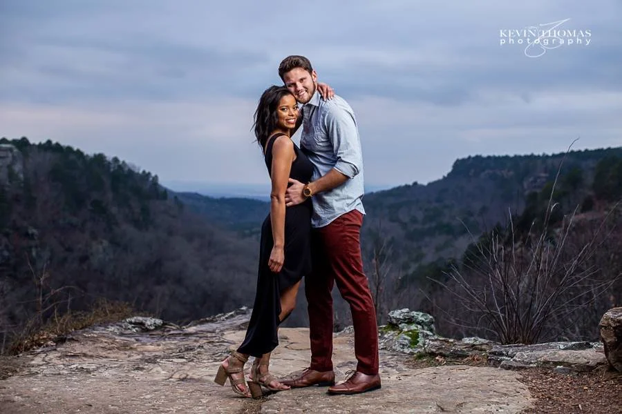 A couple embracing each other on a rocky overlook with a valley and hills in the background, under a cloudy sky.
