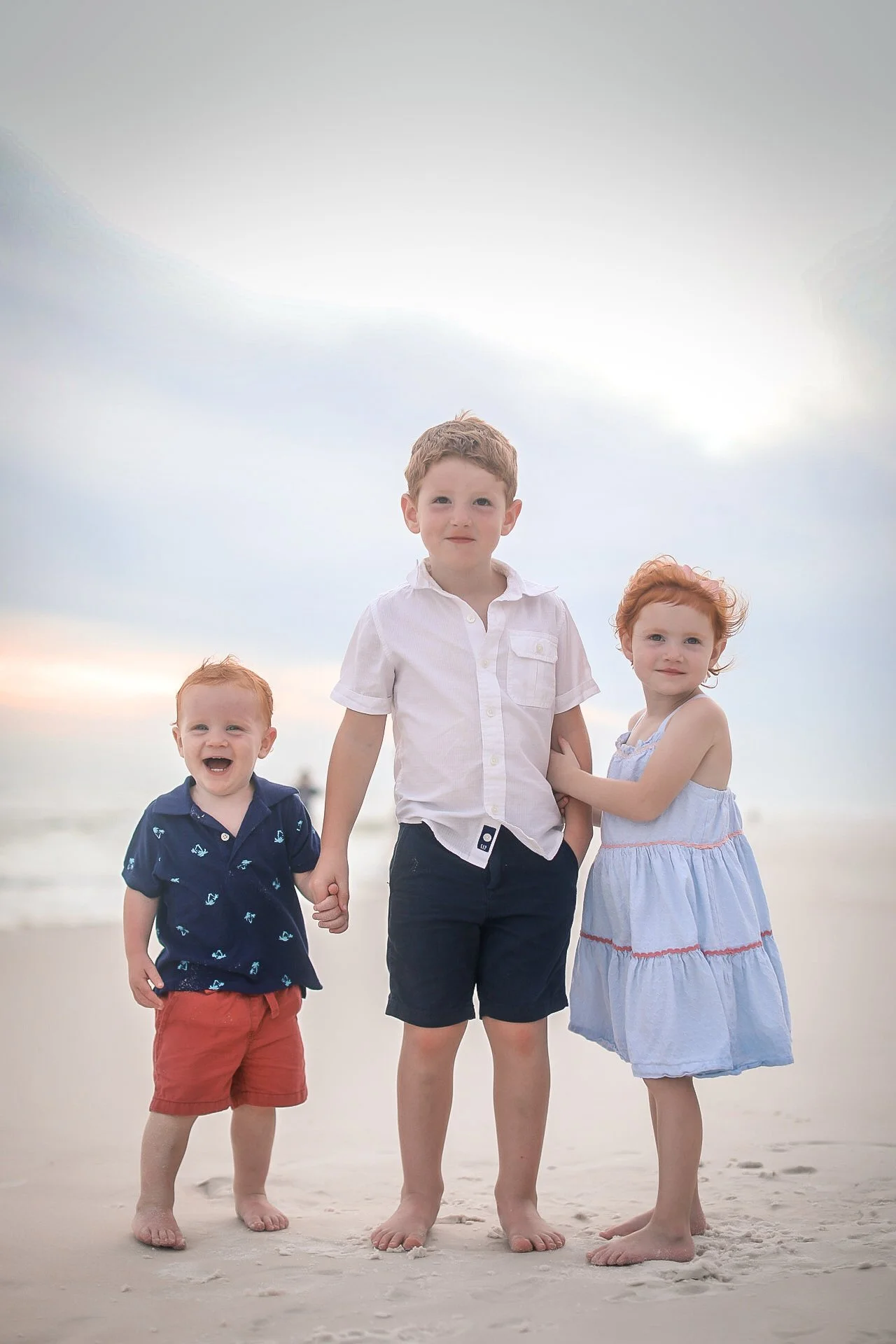Three children holding hands on a beach, with the ocean and gray sky in the background.