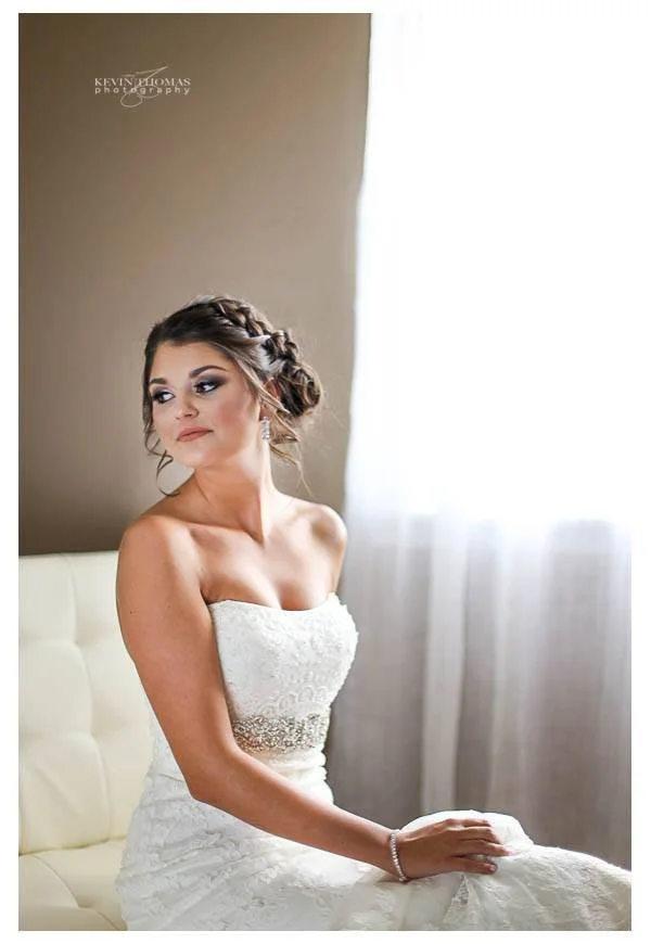 A bride in a white wedding dress sitting on a cream-colored couch by a window with sheer curtains, looking thoughtfully to the side.