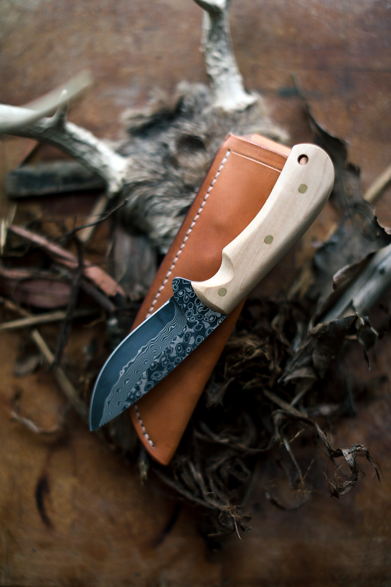 A Damascus steel hunting knife with a light wood handle and brown leather sheath, placed on leaves and twigs, with animal antlers and fur in the background.