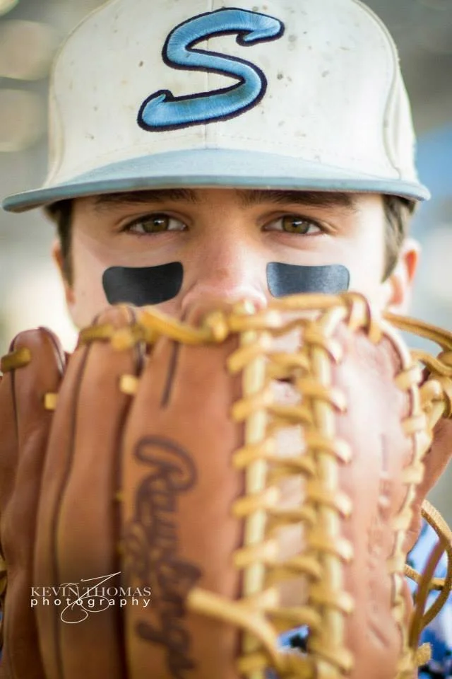 Close-up of a young baseball player wearing a white and blue cap with a raised letter 'S', black eye black under eyes, peering over a tan baseball glove.