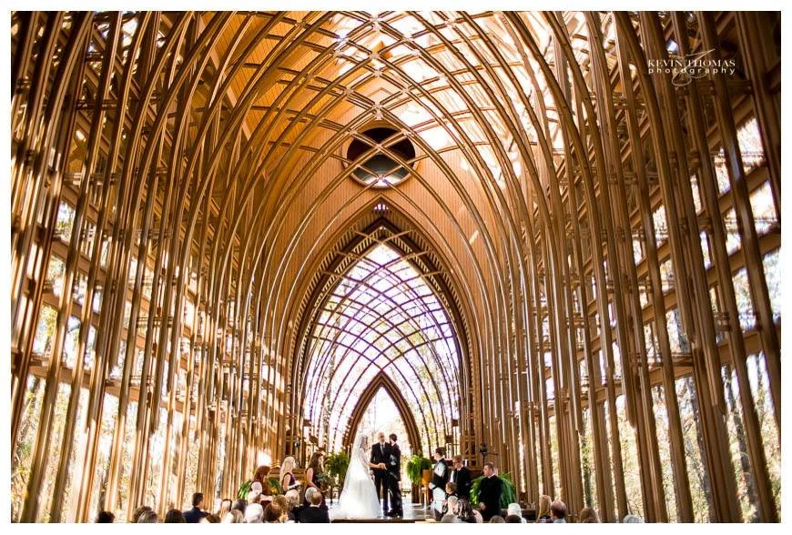 A wedding ceremony taking place inside a large, ornate glass and metal chapel with arched ceilings.