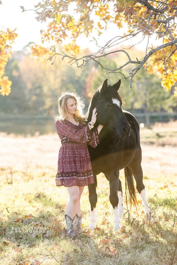 A young woman stands next to a black and white horse in a field during autumn, with trees and falling leaves in the background.