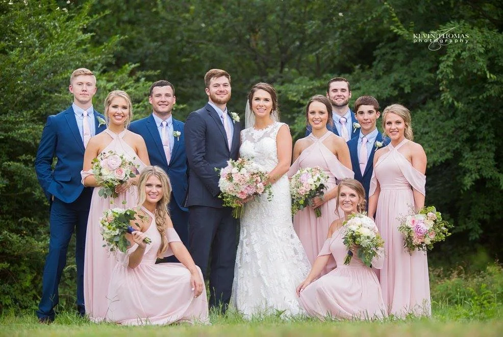 A wedding party outdoors, consisting of the bride and groom, four bridesmaids, and four groomsmen, posing on grass with green trees in the background.