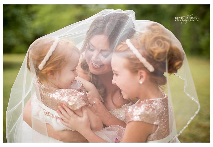 A woman with two young girls under a bridal veil, smiling and sharing a joyful moment outdoors on a grassy field.
