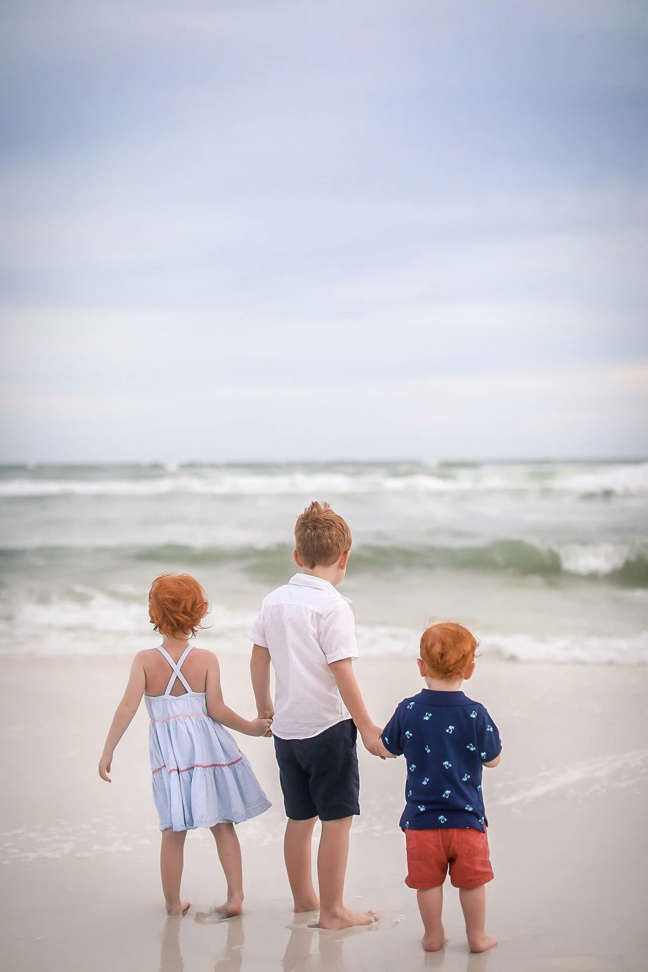 Three children, two boys and one girl, standing on the beach holding hands and looking at the ocean, in a cloudy and slightly overcast setting.