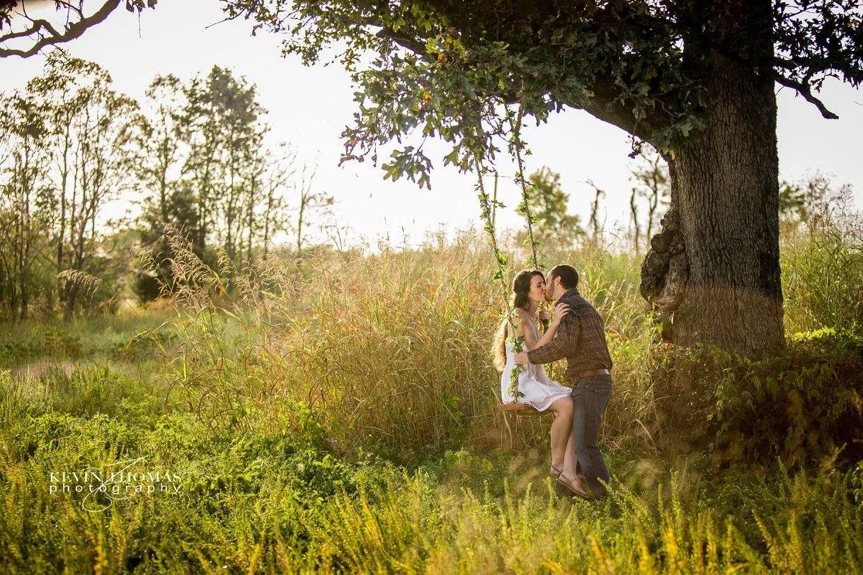 A couple kissing under a large tree in a grassy field during sunset.