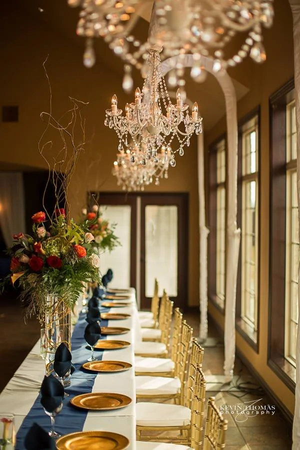 Elegant dining table setup with a long white tablecloth, gold charger plates, navy napkins, and gold chairs in a brightly lit room with large windows, floral centerpiece, and crystal chandeliers.