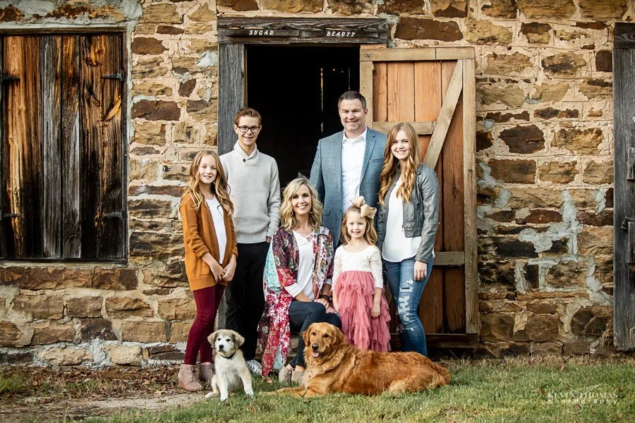 A family of seven, including a man, a woman, three girls, and two boys, standing in front of a rustic stone barn with wooden doors, along with two dogs sitting on the grass.