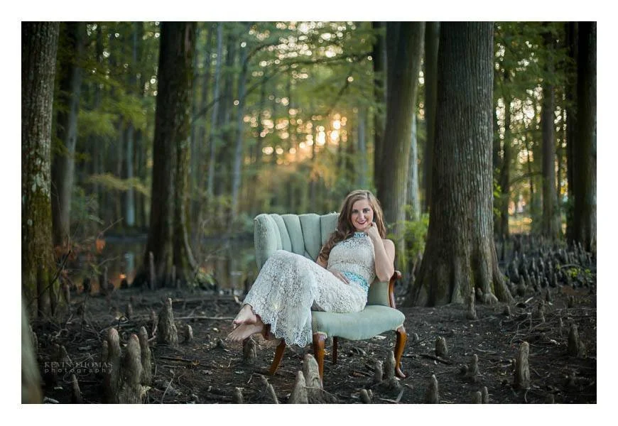 A woman in a white lace dress sitting on a vintage green armchair in a forest with tall trees and a sunset in the background.
