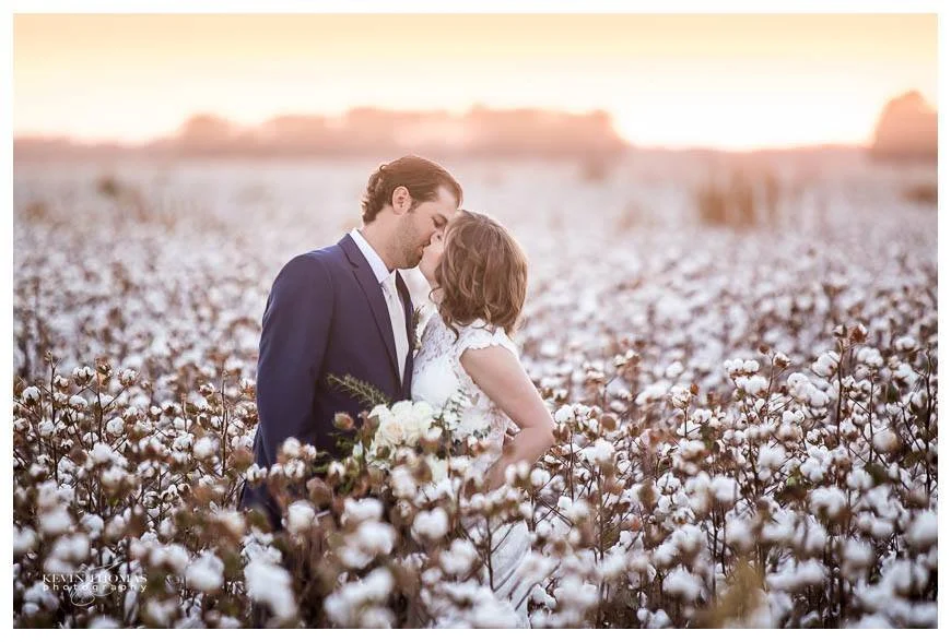 A couple dressed in wedding attire standing in a cotton field at sunset, embracing and touching foreheads.