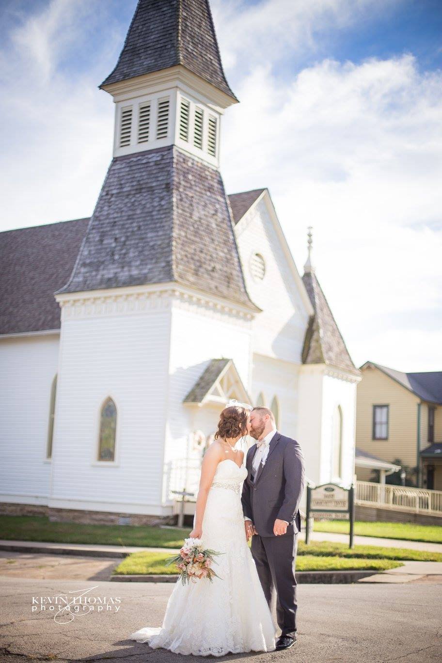 A bride and groom sharing a kiss in front of a white church with a tall steeple, during a sunny day.
