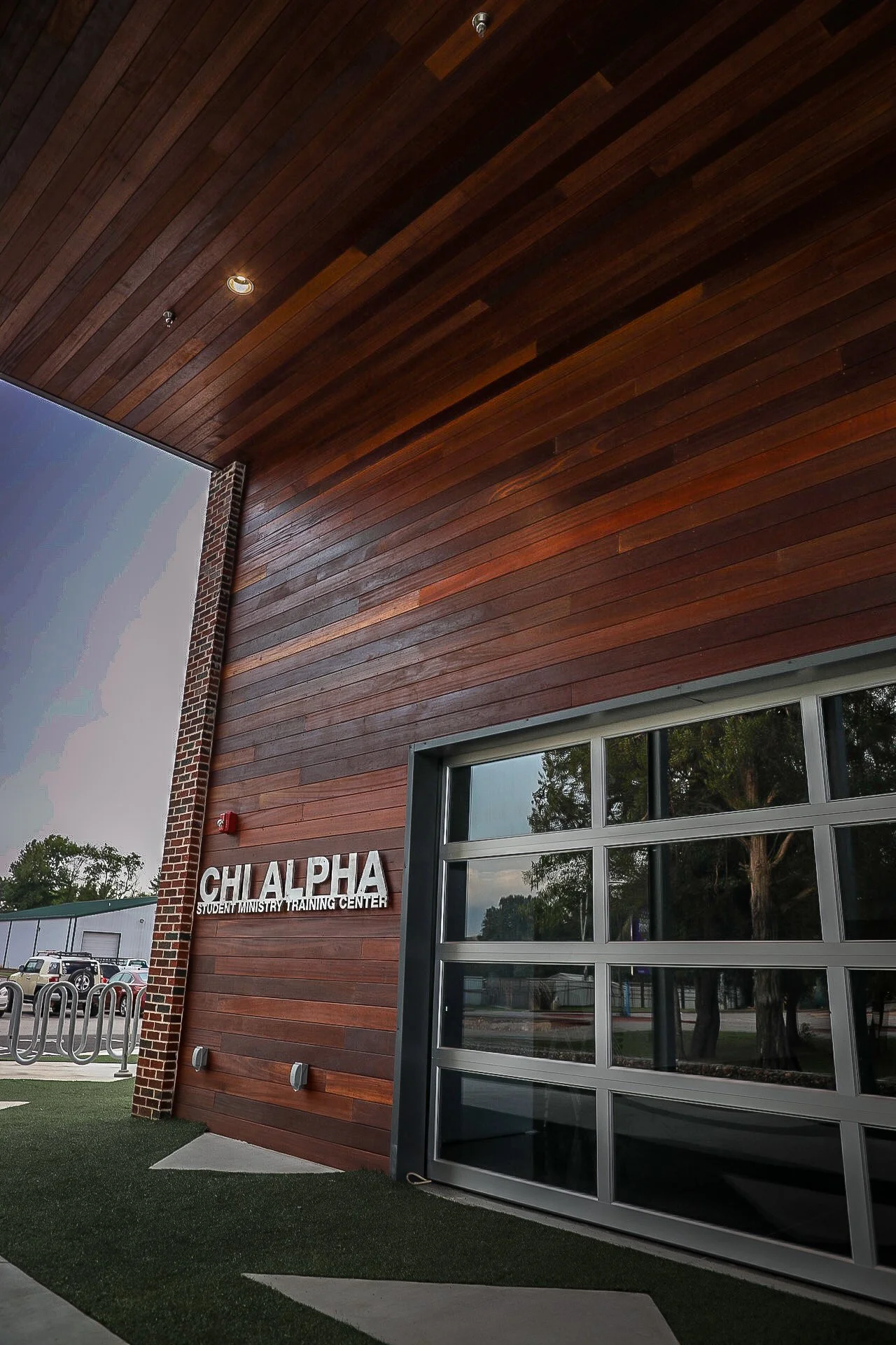 Exterior of Chi Alpha Student Ministry Training Center with a brick and wooden facade, metal garage-style door, and outdoor lighting.