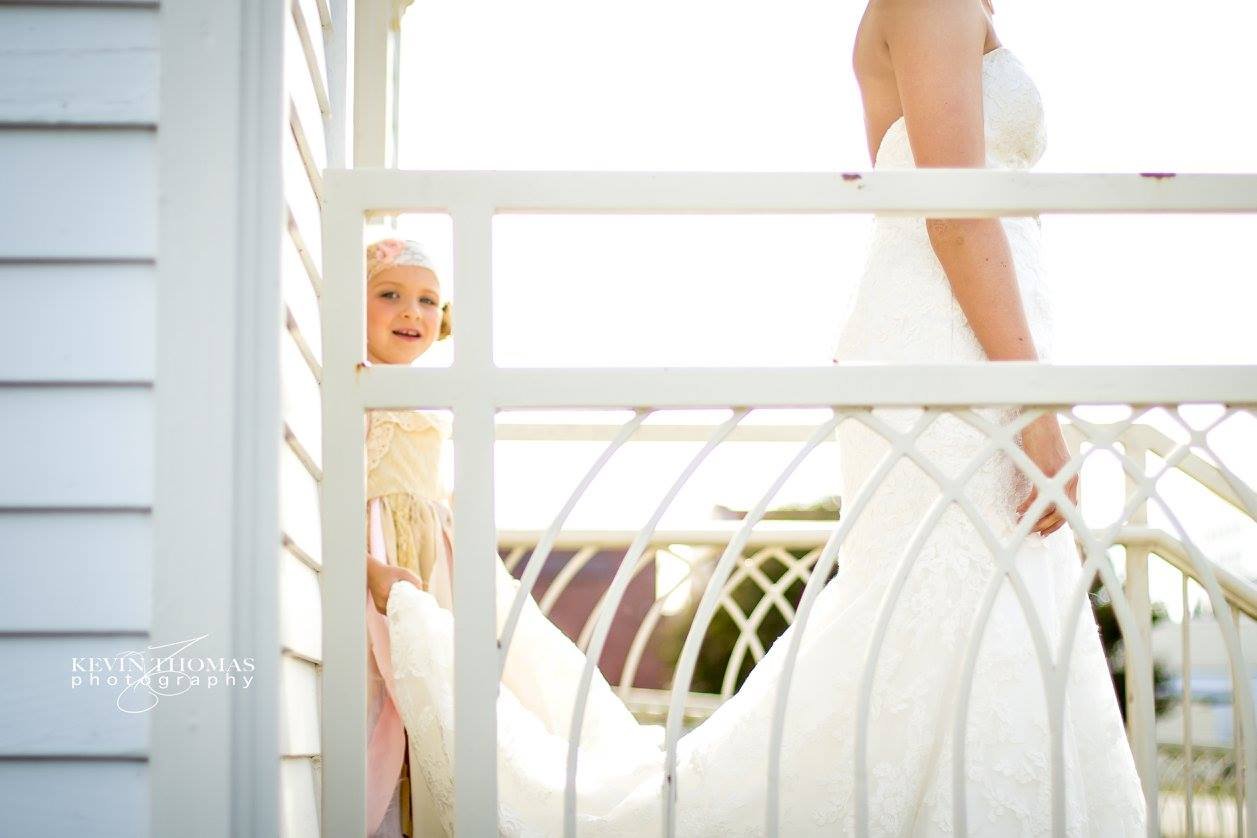 A bride in a white wedding dress standing on a balcony with a young girl in a pastel dress holding her gown.