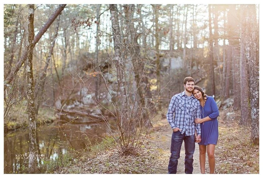 A couple standing together on a wooded path near a small body of water in a forest, holding hands with the woman resting her head on the man's shoulder, during late afternoon sunlight.