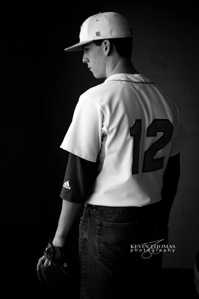 Black and white photo of a young man in a baseball jersey with the number 12, wearing a cap, and holding a glove, standing against a dark background.