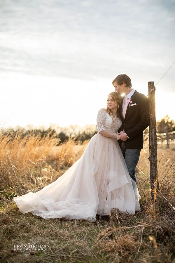 A newlywed couple stands close together in a field with tall grasses, holding hands and leaning against a wooden post as the sun sets in the background, casting a warm glow.