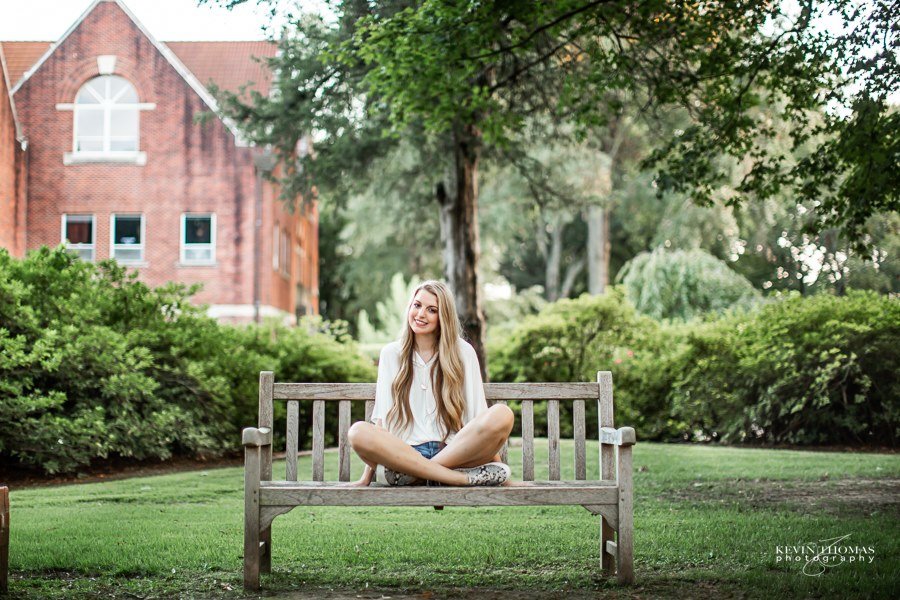 A young woman with long blonde hair, wearing a white long-sleeve shirt, shorts, and sneakers, sitting cross-legged on a wooden park bench surrounded by green grass and bushes, with a large brick building and trees in the background.